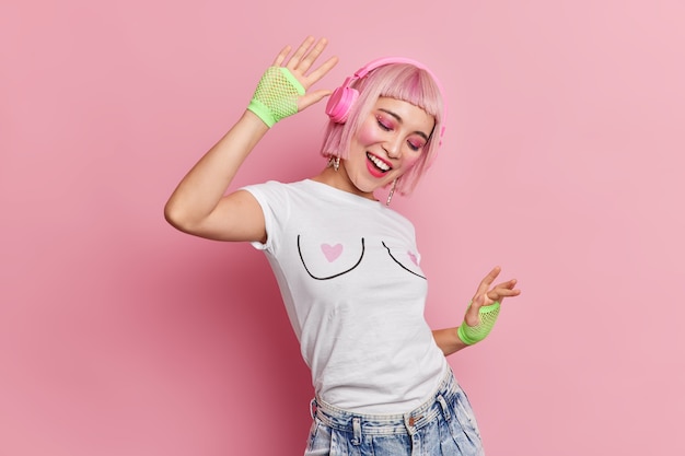 Positive millennial girl with pink hair enjoys listening music via headphones dances with rhythm of music has professional makeup