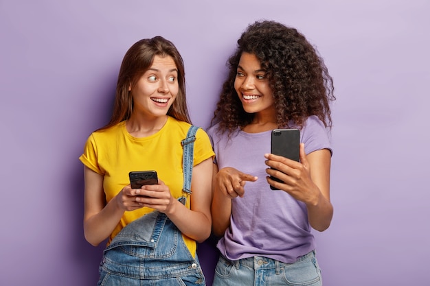 Positive millennial mixed race sisters pose with modern smartphone devices, addicted to technologies, chat online