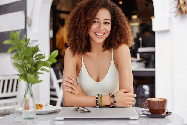 Positive mixed race female with dark skin and shining smile, enjoys coffee break, sits against cafe interior.