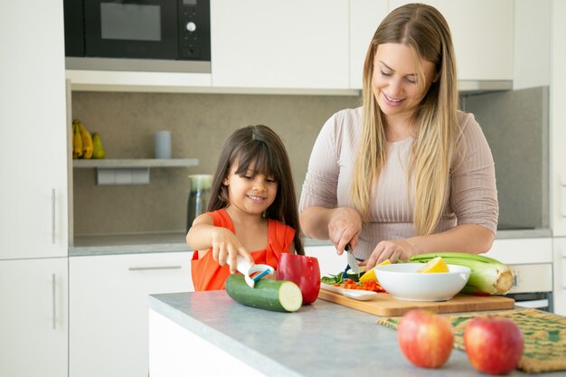 Positive mom and daughter cooking vegetables for dinner, smiling and talking. Girl and her mother peeling and cutting vegs for salad on kitchen counter. Family cooking concept