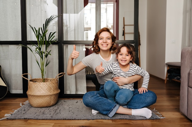 Foto gratuita la mamma positiva e la sua piccola figlia impertinente si siedono sul tappeto e mostrano i pollici in su.