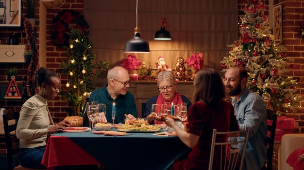 긍정적인 다문화 가족 구성원은 집에서 특별한 저녁 식사를 하며 크리스마스를 축하합니다. 함께 즐거운 시간을 보내면서 전통적인 가정 요리를 즐기는 행복한 사람들. 삼각대 촬영