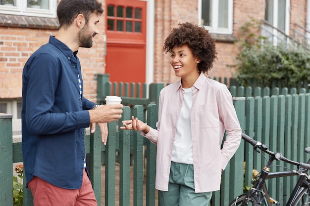 Positive multiracial couple walk in rural setting, stroll during weekends, drink takeaway coffee, stand near fence, have pleasant talk with each other