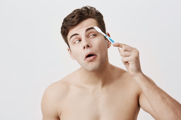 Positive naked muscular guy with trendy hairstyle, healthy skin, making faces, concentrated on combing his eyebrows with toothbrush. Attractive good-looking male posing .