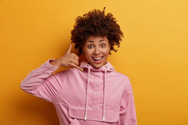 Positive optimistic curly haired millennial woman makes phone gesture of calling, call me back sign, asks for telephone number, giggles happily, wears casual hoodie. Connection communication concept