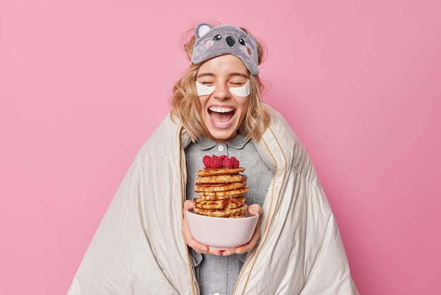 Positive optimistic woman laughs gladfully wrapped in blanket holds deicious pancakes with raspberry wears sleepmask on forehead applies beauty patches under eyes isoated over pink studiowall