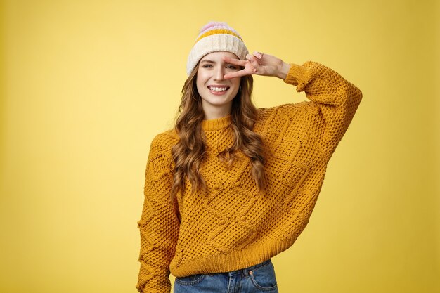 Positive peaceful kind charming young woman show peace victory gesture near eye smiling broadly have good mood perfect day wishing you good luck, standing cheerful grinning yellow background