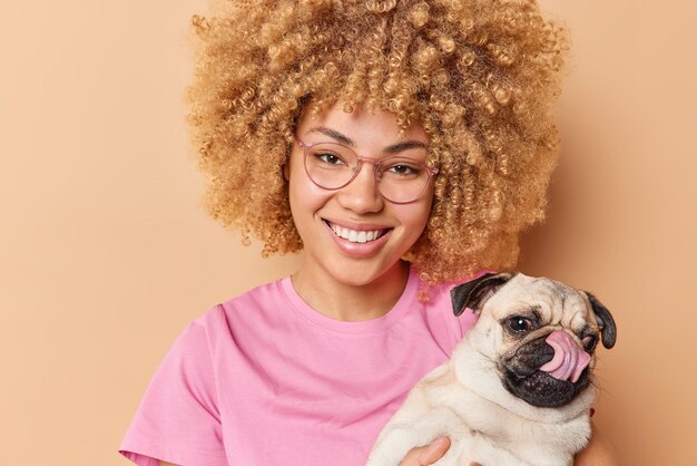 긍정적인 애완동물 주인은 캐주얼한 분홍색 티셔츠를 입고 산책을 할 퍼그 개가 포즈를 취하고 갈색 배경 위에 고립되어 기쁘게 생각합니다. 곱슬머리 여성 모델은 가축을 좋아합니다.