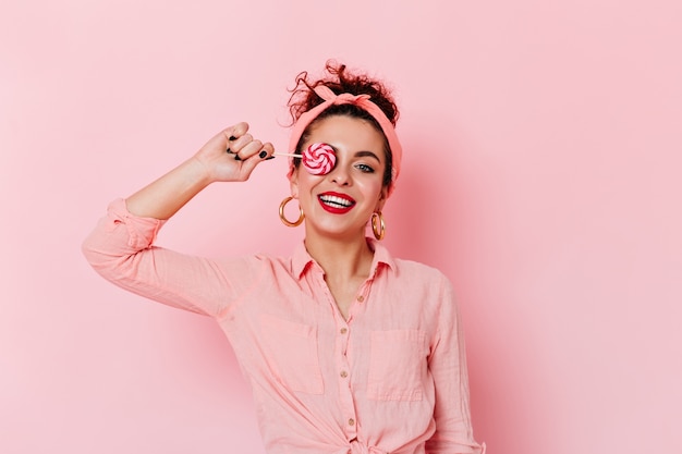 Positive pin-up girl with red lipstick in pink outfit and gold earrings holding lollipop.