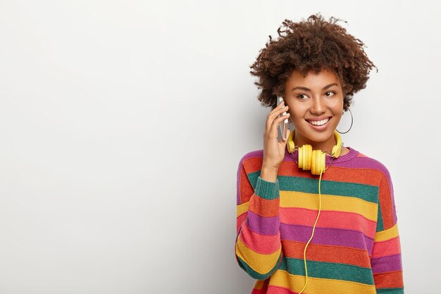 긍정적 인 유쾌한 곱슬 여성은 캐주얼 한 전화 통화를하고 캐주얼 한 복장을 입고 의사 소통을 즐기고 오디오 트랙을 듣기 위해 헤드폰을 사용합니다.