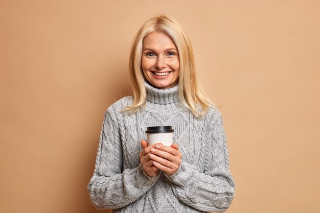Positive pleasant looking woman with blonde hair holds disposable cup of coffee enjoys drinking hot beverage during cold winter weather dressed in knitted grey sweater.