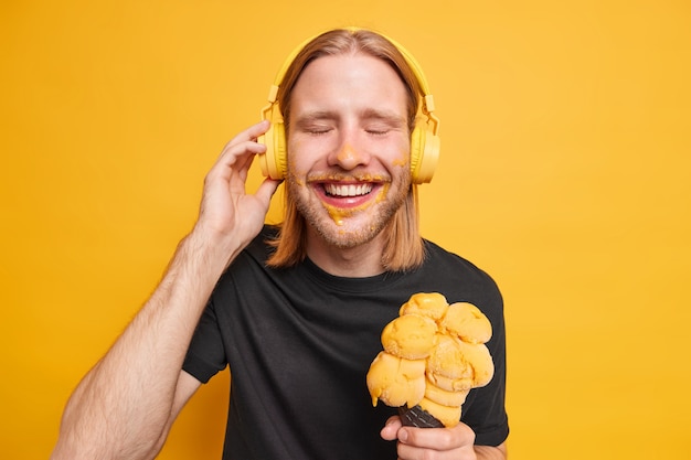 Positive pleased redhead guy closes eyes and smiles happily enjoys favorite song keeps hand on headphones holds big tasty ice cream has spare timeduring day off in summer isolated on yellow wall