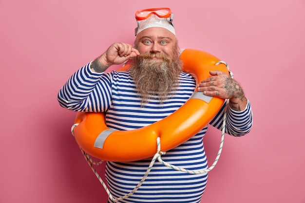 Positive plump man curls mustache wears swimming goggles and striped sailor shirt, poses with safety equipment on beach, enjoys summer holidays. Rest and season concept