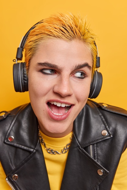 Positive punk girl with short yellow hair dark makeup dressed in fashionable clothes listens music in wireless headphones enjoys auidio podcast for entertainment