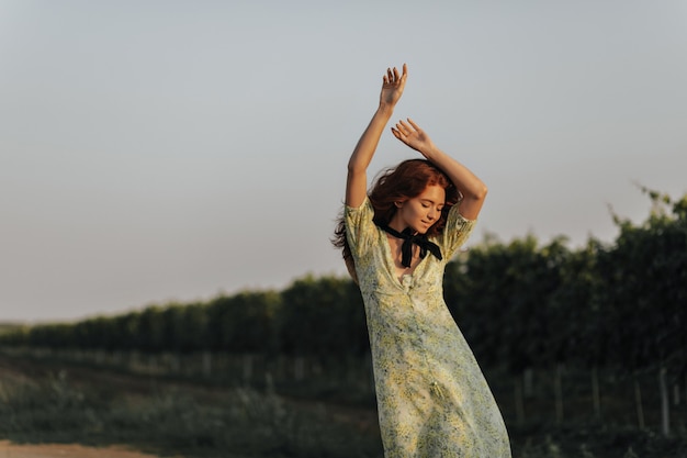 Foto gratuita signora dai capelli rossi positiva con benda nera sul collo in abiti estivi verdi che sorride e posa con le mani in alto sui vigneti