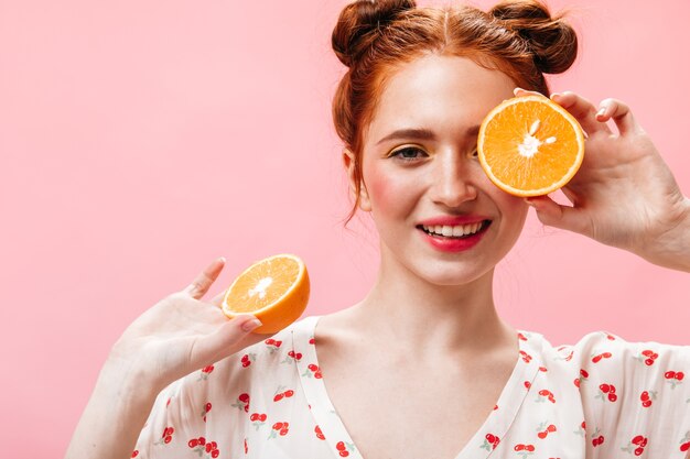 Positive red-haired woman in white dress eats juicy orange on pink background.