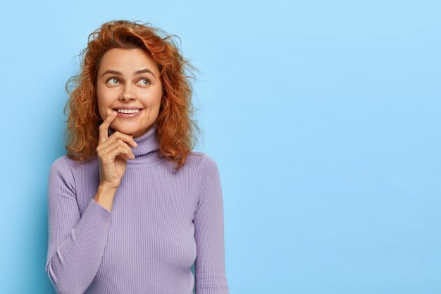 Positive redhaired woman imagines pleasant moment with boyfriend, smiles gently, looks above with dreamy face, has short red hair, wears purple sweater, isolated on blue wall, blank space for text