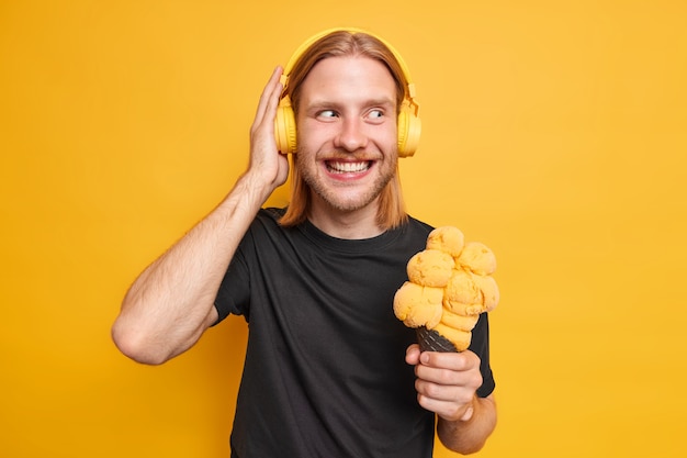 Позитивный рыжий бородатый мужчина держит руку на наушниках, держит вкусное мороженое, наслаждается любимой музыкой в наушниках, одетых небрежно изолированно над желтой стеной. Хипстерский парень с мороженым