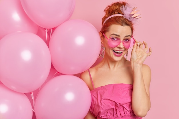 Positive redhead European girl smiles positively keeps hand on rosy sunglasses dressed in festive dress holds inflated balloons enjoys being on party celebrates graduation isolated on pink wall