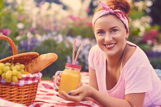 긍정적인 빨간 머리 여성은 과일, 빵, 와인으로 가득 찬 피크닉 바구니와 함께 벤치에 앉아 있습니다.