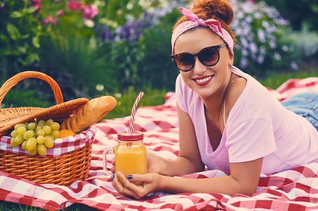 Positive redhead female sits on a bench with a picnic basket full of fruits, bread and wine.