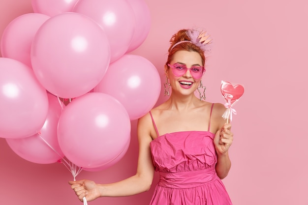 Positive redhead girl in trendy pink shades and dress holds tasty sweet candy and bunch of balloons has festive mood on party poses against rosy background.