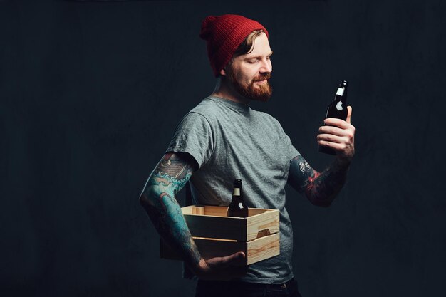 Positive redhead tattooed, bearded male holds a wooden box with beer bottles over grey background.