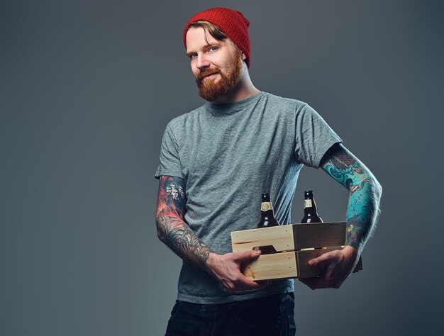 Позитивный рыжий татуированный бородатый мужчина держит деревянную коробку с пивными бутылками на сером фоне.