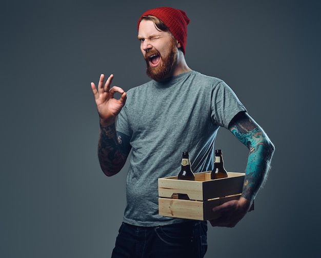 Позитивный рыжий татуированный бородатый мужчина держит деревянную коробку с пивными бутылками на сером фоне.
