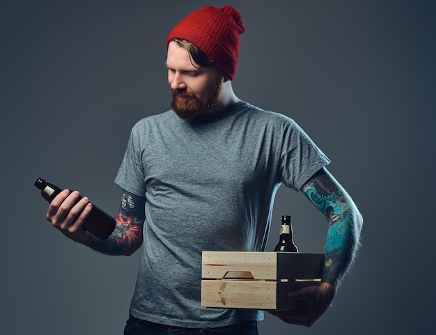 Positive redhead tattooed, bearded male holds a wooden box with beer bottles over grey background.