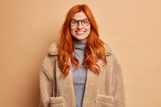 Positive redhead woman dressed in warm fur coat smiles pleasantly has good mood expresses happy emotions.