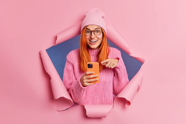 현대 스마트 폰 디스플레이의 긍정적 인 빨간 머리 여성 포인트는 소셜 네트워크의 인터넷 서핑에서 유용한 정보를 얻습니다. 웃음은 모바일 앱이나 신기술을 적극적으로 사용합니다. Blogger 라이프 스타일.