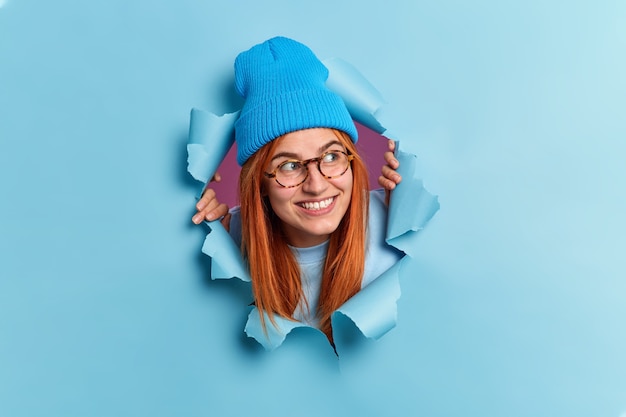Positive redhead young woman looks away with pleasant smile has curious expression wears hat and optical glasses breaks through blue paper