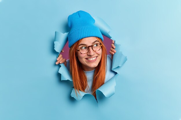 Positive redhead young woman looks away with pleasant smile has curious expression wears hat and optical glasses breaks through blue paper