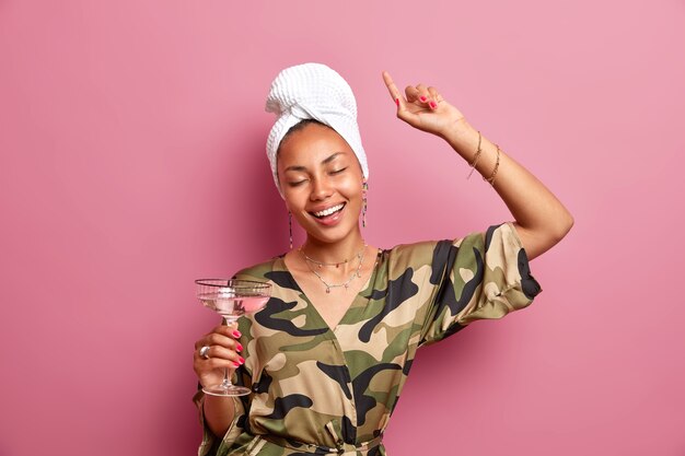 Positive relaxed ethnic woman with dark skin keeps eyes closed raises arm poses with cocktail at home has fun at domestic party wears khaki dressing gown white towel wrapped on head models indoor