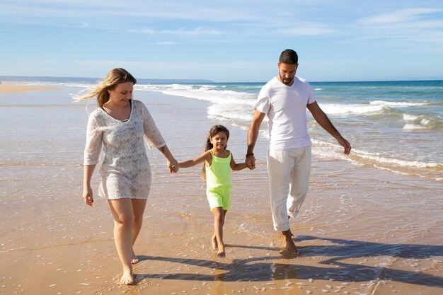 Позитивная расслабленная семейная пара и маленькая девочка гуляют по мокрому золотому песку на пляже, ребенок держится за руки родителей