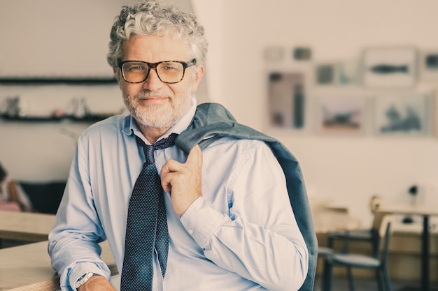 Positive relaxed mature business man standing in office cafe, leaning on counter, holding jacket over shoulder