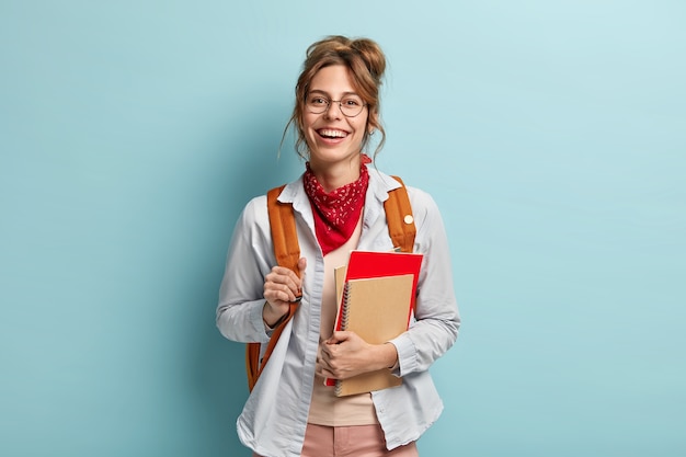 Positive schoolgirl carries spiral notepad, notebook, carries backpack, ready for school and lessons