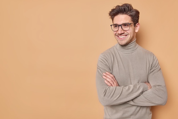 Positive self confident man keeps arms folded looks away with satisfied expression listens good news wears spectaces and casual turtleneck isolated over beige background blank space for promo
