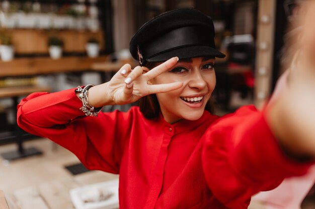 Positive selfie of bright smiling young lady with beautiful eyes, good manicure, gorgeous makeup in a beautiful silk dress with black belt. Model shows peace sign with her fingers