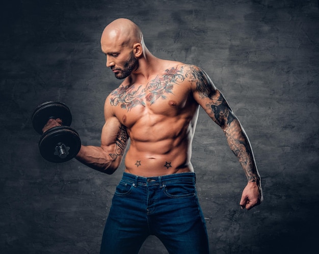 Free photo positive shaved head shirtless male dressed in a jeans holds the dumbbell over dark grey background.