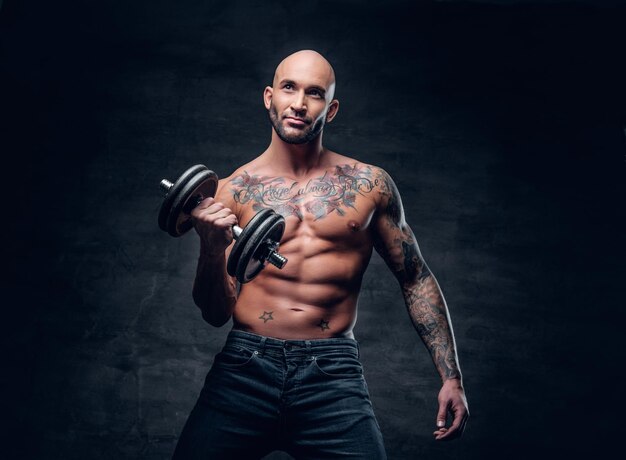 Positive shaved head shirtless male dressed in a jeans holds the dumbbell over dark grey background.