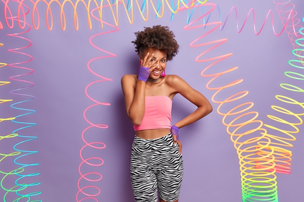 Positive shy curly haired woman makes face palm, wears tank top, leggins and sport gloves, being in good mood