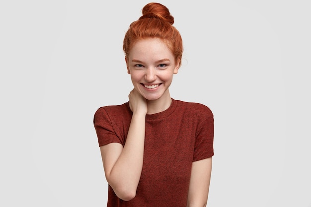 Positive shy ginger girlfriend has tender smile, keeps hand on neck, being in high spirit