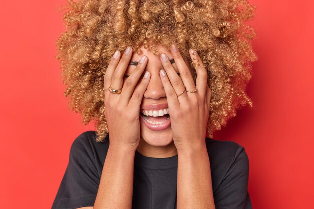 Positive sincere woman covers eyes with hands laughs happily has upbeat mood smiles gladfully dressed in casual black t shirt isolated over vivid red background. authentic emotions and feelings
