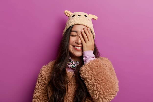 Free photo positive sincere young woman laughs with joy, covers half of face, keeps eyes closed, dressed in winter outerwear