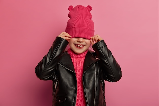 무료 사진 긍정적 인 작은 아이는 분홍색 모자로 얼굴을 숨기고, 눈을 가리고, 가죽 자켓을 입고, 장난스런 행복한 미소를 지으며, 장밋빛 벽에 포즈를 취하고, 낙관적이며, 세련된 복장을 시도합니다. 어린이 개념