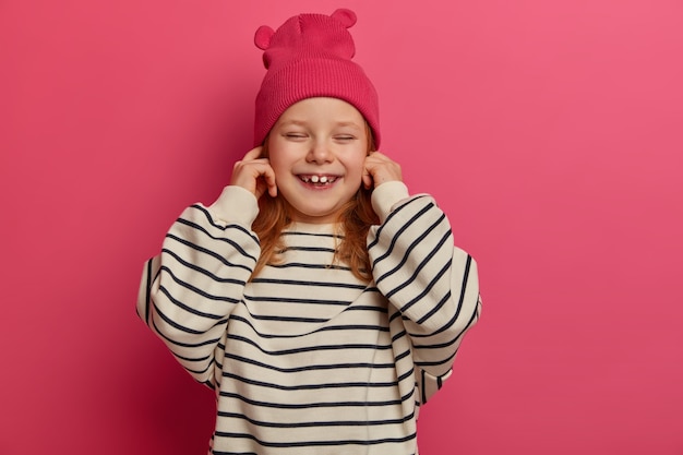 Positive small child plugs ears, ignores loud sound, closes eyes and chuckles, wears pink hat and oversized striped jumper, poses indoor. Little girl being naughty, doesnt want to hear parents