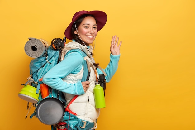 Positive smiling Asian woman packpacker has cheerful expression, waves palm at camera, carries all necessary things in big rucksack