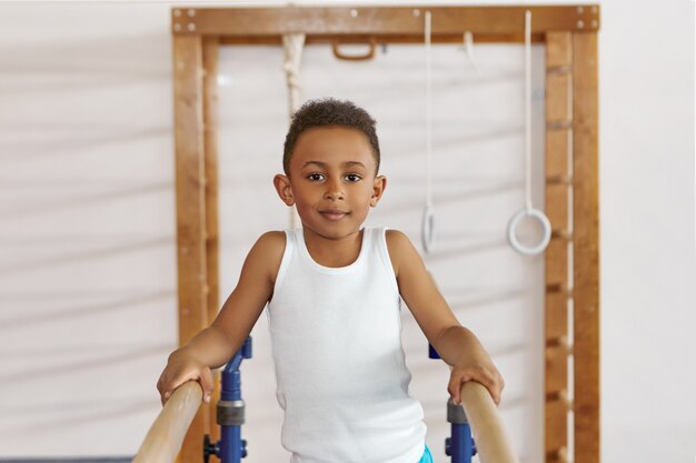 Positive smiling black dark skinned boy in white tank top exercising on two wooden parallel bars